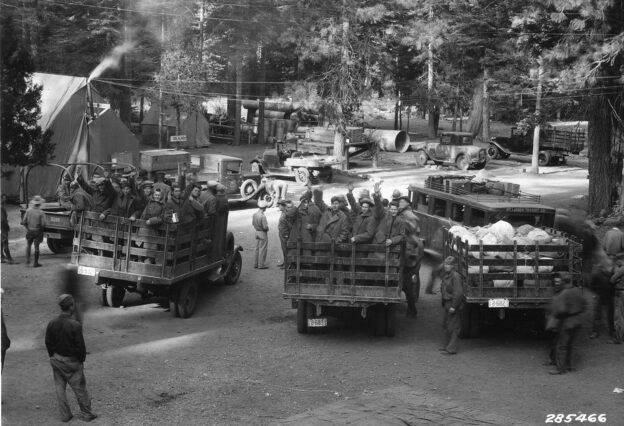 Three trucks, two of them carrying enrollees from the Civilian Conservation Corps as they get ready to leave their camp and conclude their term of service. Trees and tents are visible behind the trucks: the camp is in a forest. Some men look on.